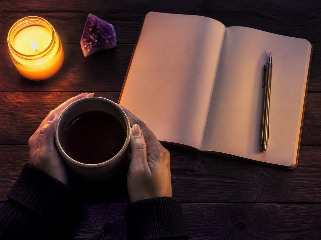 Hands holding a warm cup beside an open journal and a candle